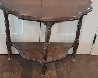May include: A dark brown wooden half-moon console table featuring a semi-circular top with a scalloped edge. The table is supported by three turned legs and includes a lower semi-circular shelf, showcasing a rich wood grain finish throughout.