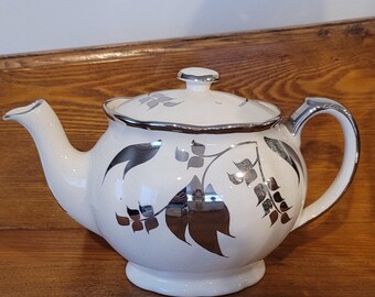 May include: White ceramic teapot with a silver rim, handle, and spout. The teapot features a floral design in silver. The lid has a small silver knob. The teapot sits on a wooden surface.