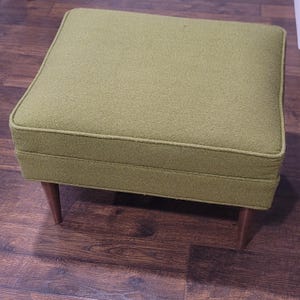 May include: A square ottoman upholstered in a textured olive green fabric with piped edges. The piece features four tapered wooden legs that extend outward from the base, providing a stable foundation for the cushioned seat top.