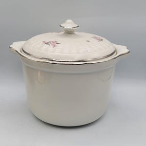 May include: A white ceramic casserole dish with a lid. The lid features a floral pattern with pink flowers and a silver trim. The dish has a silver trim and handles on either side. The casserole dish is a kitchenware item.