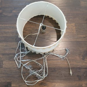 May include: A round, off-white fabric lampshade with a decorative scalloped fringe trim along the bottom edge. The shade features a central metal spider fitter with a single light socket, suspended by a silver-toned metal chain and power cord.