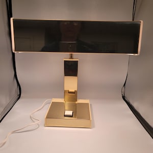 Puede incluir: Una lámpara de escritorio vintage con una pantalla rectangular negra y una base de metal dorado. La lámpara tiene una base rectangular con un interruptor de encendido/apagado blanco. La lámpara está conectada a un cable eléctrico blanco.