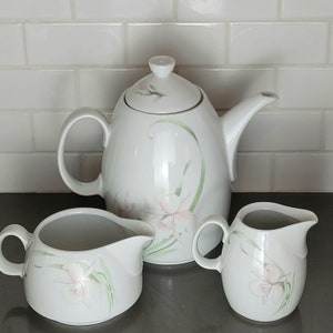 Könnte beinhalten: Eine weiße Porzellan-Teekanne und zwei Milchkännchen mit einem Blumendesign. Die Teekanne hat einen Deckel und einen Ausguss. Die Milchkännchen haben einen Griff und einen Ausguss. Das Blumendesign ist in Rosa-, Grün- und Weißtönen gehalten.