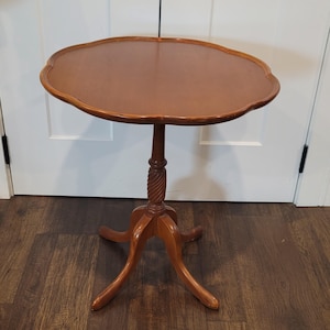 Peut inclure: Une table d'appoint ronde en bois avec un plateau à bord festonné. La table présente un pied central avec un détail de colonne sculptée en spirale et trois pieds incurvés s'étendant vers l'extérieur pour assurer la stabilité du plateau.