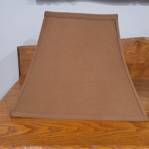 Puede incluir: Una pantalla de lámpara marrón con forma trapezoidal rectangular. La pantalla está hecha de una tela marrón claro lisa y tiene un ribete cosido a lo largo de los bordes superior e inferior. La pantalla está sobre una superficie de madera.