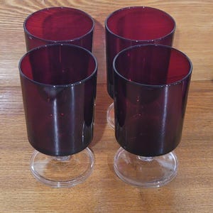 May include: Four ruby red stemmed glasses. The glasses have a cylindrical shape with a clear, circular base. The red glass contrasts with the clear base. The glasses are arranged on a wooden surface.