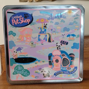 Puede incluir: Una lata metálica cuadrada con una colorida ilustración de un juego de Littlest Pet Shop. El diseño incluye varias figuras de animales, edificios y un paisaje con cielo azul y sol. La lata está etiquetada como "Littlest Pet Shop" y "Collector's Edition".