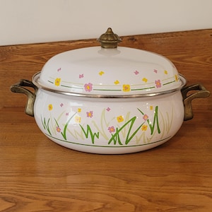 May include: White enamel cooking pot with a lid, decorated with pink and yellow flowers and green grass. The pot has brass-coloured handles and a decorative knob. The pot is on a wooden surface. A charming kitchenware item.