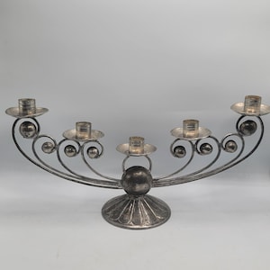 Peut inclure: Un candélabre en métal argenté doté d'une base large et incurvée avec cinq bougeoirs. La structure est ornée de sphères métalliques décoratives et d'accents en fil torsadé, reposant sur un pied circulaire nervuré pour plus de stabilité.
