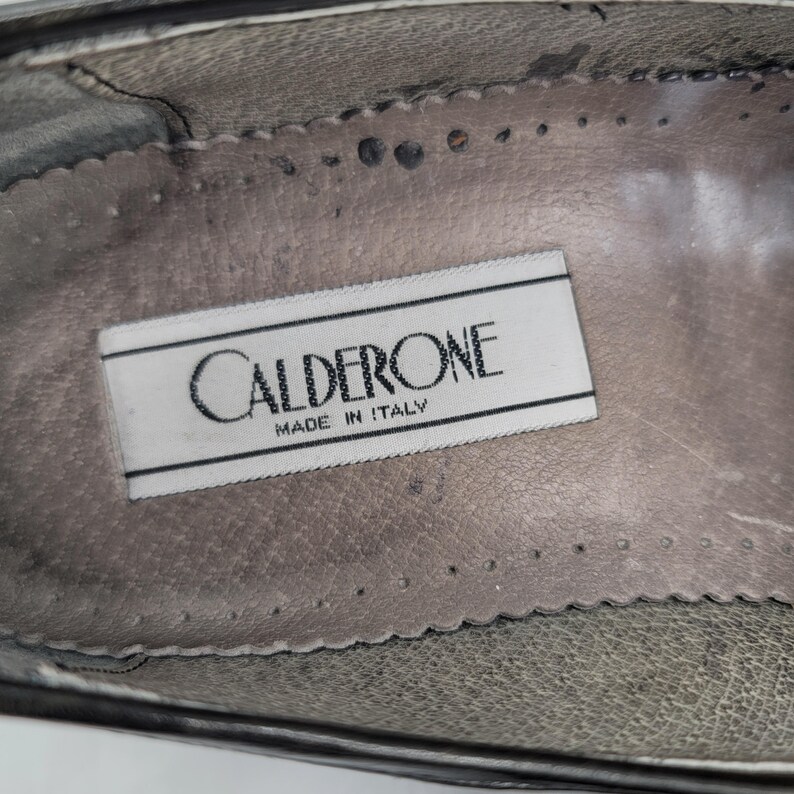 May include: Close-up of the inside of a shoe, featuring a label that reads "CALDERONE MADE IN ITALY" in black text on a white rectangular tag. The shoe's interior is a worn, textured gray color.