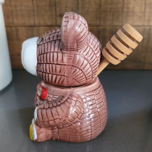 May include: A brown ceramic honey pot shaped like a bear with a wooden honey dipper. The bear has a textured surface and a white face.