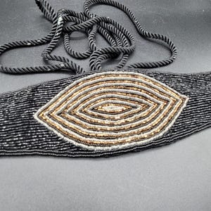 May include: A black beaded belt featuring an oval gold and silver beaded design. The belt has a black rope tie. This fashion accessory is suitable for various outfits.