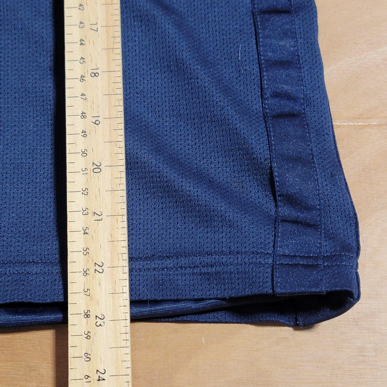 Puede incluir: Primer plano del borde inferior de una camiseta deportiva azul marino con una costura blanca en el lateral. Se puede ver una regla de madera en la imagen, que muestra que la longitud de la camiseta es de 58,4 cm.