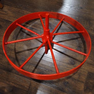 Könnte beinhalten: Ein hellrotes Metallrad mit Speichen, wahrscheinlich von einem Wagen oder Karren. Das Rad hat eine zentrale Nabe und einen kreisförmigen Felgenrand, wobei die Speichen nach außen strahlen. Das Rad ist einfarbig rot lackiert.