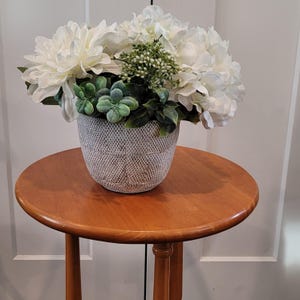 Peut inclure: Un arrangement floral avec de grandes fleurs blanches, du feuillage vert et de petits bourgeons verts dans un pot gris texturé. Le pot est posé sur une table d'appoint ronde en bois. La table a une finition brun-rougeâtre chaude.