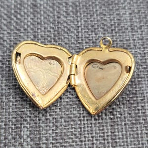 May include: A gold-toned heart-shaped locket shown in an open position. The interior features two empty compartments with a hinged centre. The left side displays a faint embossed heart pattern, while the right side has a smooth, plain surface.