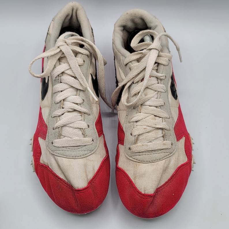 May include: A pair of white and red running shoes with black accents. The shoes have a white canvas upper with red trim around the toe and heel. The laces are white and the shoes have a black rubber sole.
