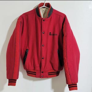 May include: A vintage red varsity jacket with black accents. The jacket features black leather pockets, a black and white striped collar and cuffs, and the text "HEV MOTORS" on the chest. The jacket is button-down and has red stripes down the sleeves.
