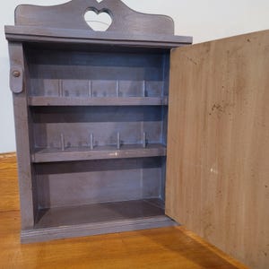 Peut inclure: Un meuble mural en bois gris avec une découpe en forme de cœur en haut. Le meuble a une porte ouverte, révélant des étagères et des chevilles à l'intérieur. Le meuble est conçu pour contenir de petits objets.