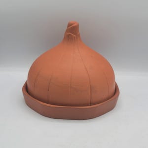 Puede incluir: Una campana de color terracota, con forma de cebolla, se asienta sobre una base a juego. La campana tiene crestas verticales y una parte superior puntiaguda. Esta pieza decorativa probablemente se utiliza para servir o exhibir alimentos.