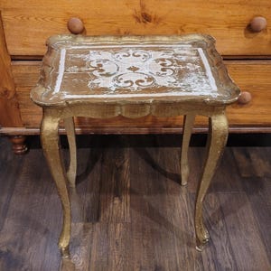 May include: A rectangular side table with a gold finish and four curved legs. The tabletop features a distressed white and gold decorative pattern with a scalloped edge. The surface shows signs of wear and aging, with visible paint texture.