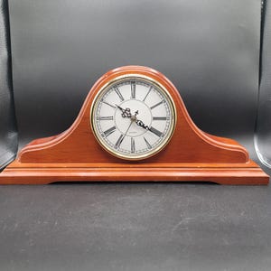 May include: A wooden mantel clock with a curved top and a rectangular base. The clock face is white with black Roman numerals and gold-coloured hands. The clock is made of reddish-brown wood.