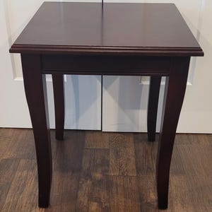 May include: A square side table with a dark brown wood finish. The table features a flat top surface with a beveled edge and four slightly curved legs that taper towards the bottom. The construction is simple with a smooth, polished surface.