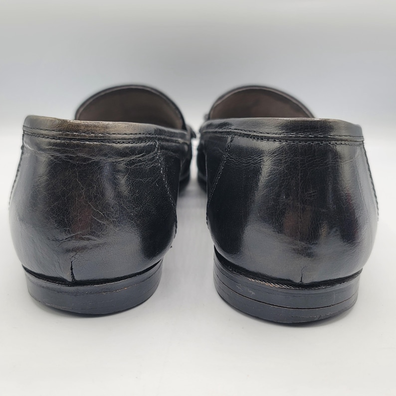 May include: Pair of black leather loafers. The shoes have a glossy finish and a classic design. The image shows the back of the shoes, highlighting the heel and sole details. The loafers appear to be well-worn.