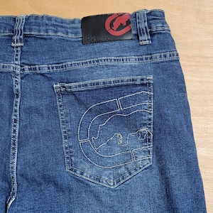 May include: Blue denim jeans with a back pocket featuring a white embroidered logo of a bear inside a circle. The logo above the pocket reads "Ecko Unltd."