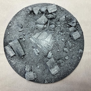 May include: A round, grey, textured base with a variety of small, grey, rectangular shapes scattered across the surface. The base is designed for miniature gaming.