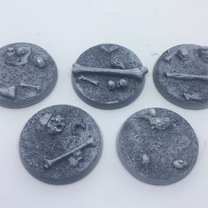 May include: Five gray plastic miniature bases with a textured surface and sculpted details of bones and skulls.