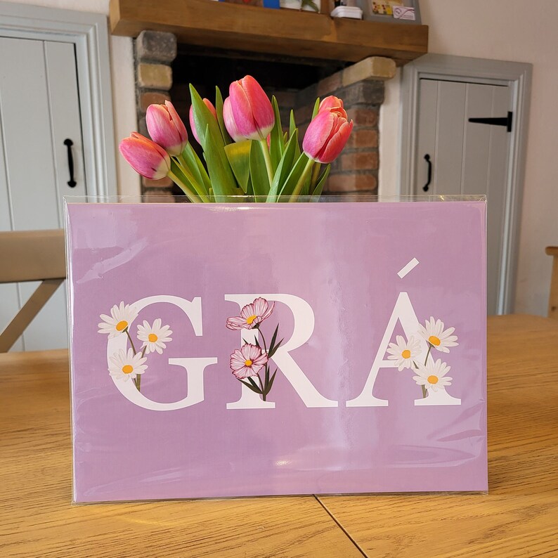 May include: A light purple print with the word "GR&Aacute;" in white letters. The letters are decorated with small white and pink flowers.