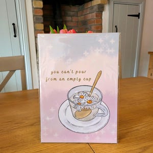Könnte beinhalten: Eine rosa und weiße Karte mit dem Zitat "Man kann nicht aus einer leeren Tasse gießen". Die Karte zeigt eine handgezeichnete Illustration einer Teetasse mit darin schwimmenden Gänseblümchen.