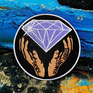 May include: A black and white embroidered patch with two hands holding a purple diamond. The hands have the words "Hold" and "Diamond Hands" written on them.