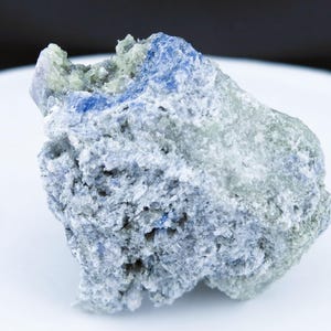 May include: A close-up of a rough, textured rock with shades of blue, gray, and green. The mineral appears crystalline and sits on a white surface. The rock has a natural, organic shape.