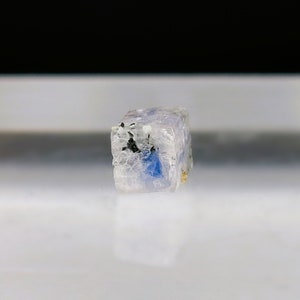 May include: A small, clear, cube-shaped crystal with blue and black inclusions. The crystal is translucent and has a slightly rough surface.