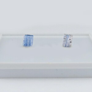 May include: Two small, rectangular, light blue kyanite crystals with a translucent, striated appearance. The stones have a natural, rough-cut texture and are positioned side-by-side on a flat, white display surface, showcasing their distinct blue hue.