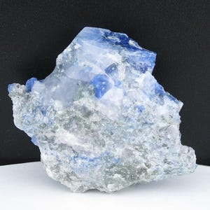 May include: A raw, natural mineral specimen featuring a mix of blue and gray hues. The crystalline structure has a rough, textured surface with areas of deep blue and lighter gray tones. The mineral sits on a white surface against a black background.