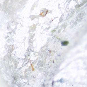 Może przedstawiać: Zbliżenie na przezroczysty, krystaliczny minerał o szorstkiej, teksturowanej powierzchni. Minerał ma przeważnie biały kolor z subtelnymi odcieniami brązu, zieleni i błękitu. Obraz podkreśla naturalny, surowy wygląd.