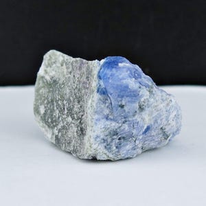 May include: A rough, natural stone with a split color design. The left side is a muted green-gray, while the right side is a vibrant blue. The stone has a textured surface and is likely a mineral specimen.