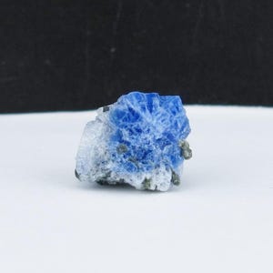 May include: A small, rough-cut mineral specimen with a vibrant blue crystalline structure. The stone has a light gray base with flecks of darker gray and sits against a white surface with a black background.