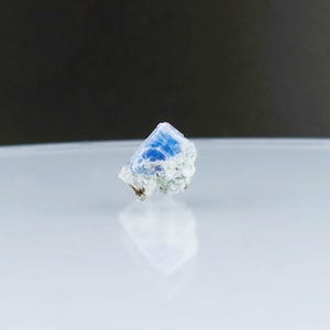 May include: A small, blue and white crystal with a rough, natural texture. The crystal is sitting on a white surface.