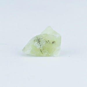 May include: A raw, pale lime-green mineral specimen with a translucent, crystalline structure. The stone features irregular, jagged edges and small, dark inclusions embedded within its surface, showcasing a natural, unpolished, and rough texture.
