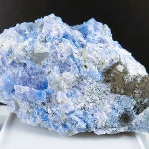 May include: A close-up of a mineral specimen with a crystalline structure. The rock features a mix of light blue, white, and gray hues, with a patch of darker, brownish-gray material. The surface has a rough, textured appearance.