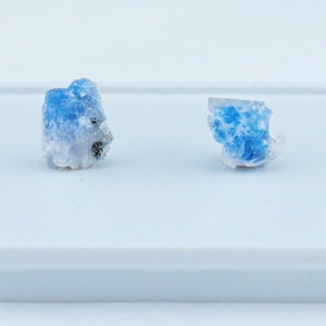 May include: Two small, raw mineral specimens featuring vibrant blue crystalline structures set against a white, translucent base. The stones exhibit irregular, jagged edges and a rough, natural texture, showcasing a mix of bright blue and white hues.