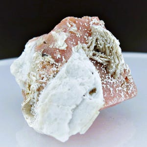 May include: A pink and white mineral specimen with a rough, textured surface. The mineral has a layered appearance with white crystals on top of a pink base.