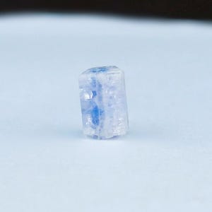 May include: A small, rectangular, translucent crystal with a light blue and white color scheme. The crystal has a rough, natural texture and is standing upright against a white background. The crystal's interior has a vibrant blue hue.
