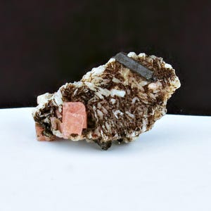 Może przedstawiać: Naturalny okaz minerału z skupiskiem kryształów. Okaz prezentuje mieszankę kolorów, w tym jasnoróżowy, brązowy i biały. Ciemnoszary, prostokątny kryształ jest osadzony na górze. Minerał leży na białej powierzchni.