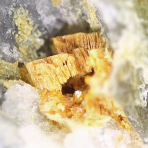 Könnte beinhalten: Nahaufnahme einer Mineralprobe mit geschichteten, goldbraunen Kristallstrukturen in einer Matrix aus grauem und weißem Gestein. Die Kristalle haben ein faseriges Aussehen, mit einer kleinen sichtbaren Höhle.