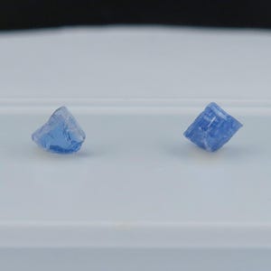 May include: Two small, faceted, blue gemstones are displayed on a white surface. The gemstones have a crystalline structure and a vibrant, light blue color. The background is a soft white, providing a clean contrast to the gems.